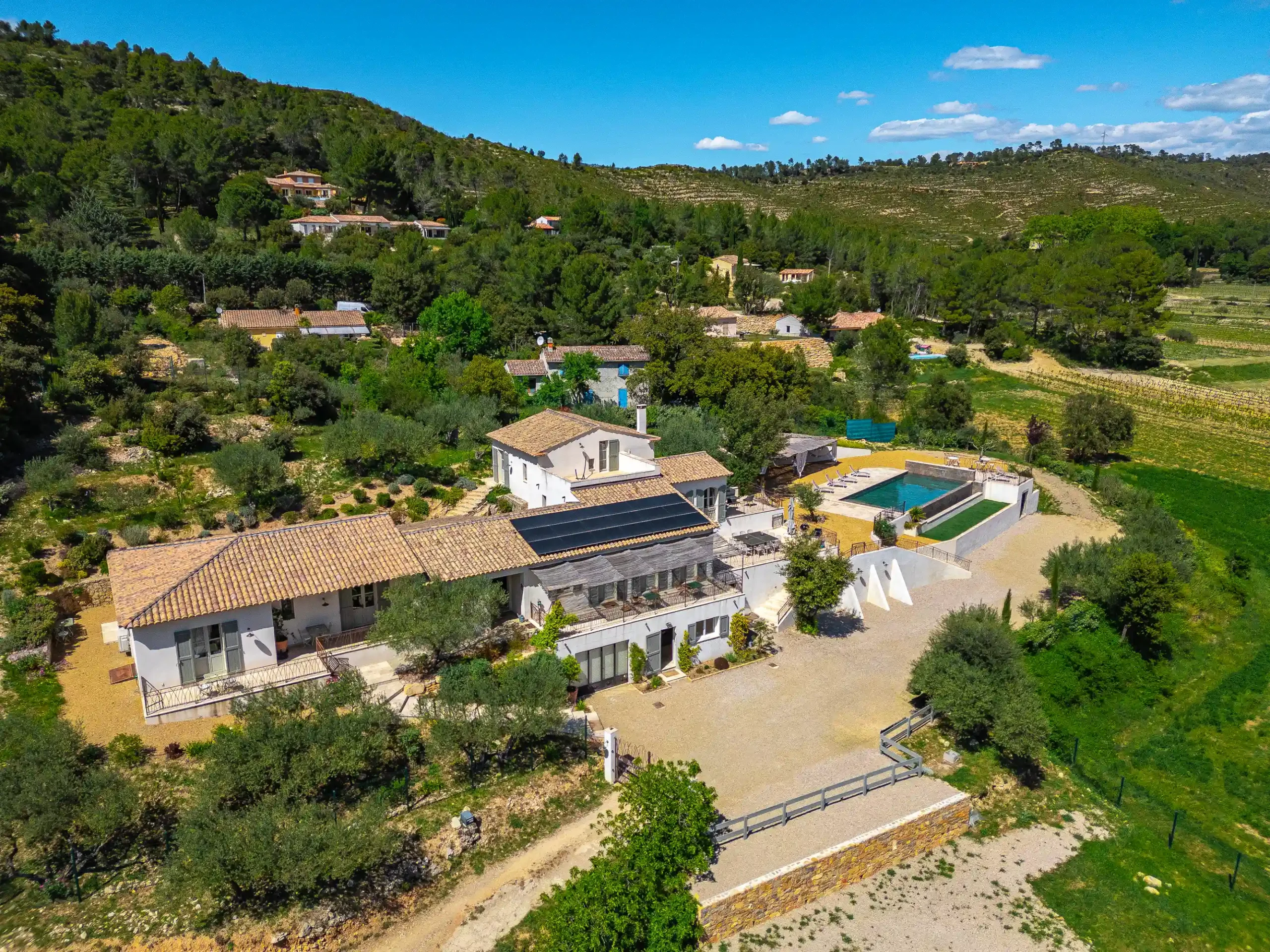 Vue aérienne de la bastide, sa piscine, les maisons voisines et les collines boisées