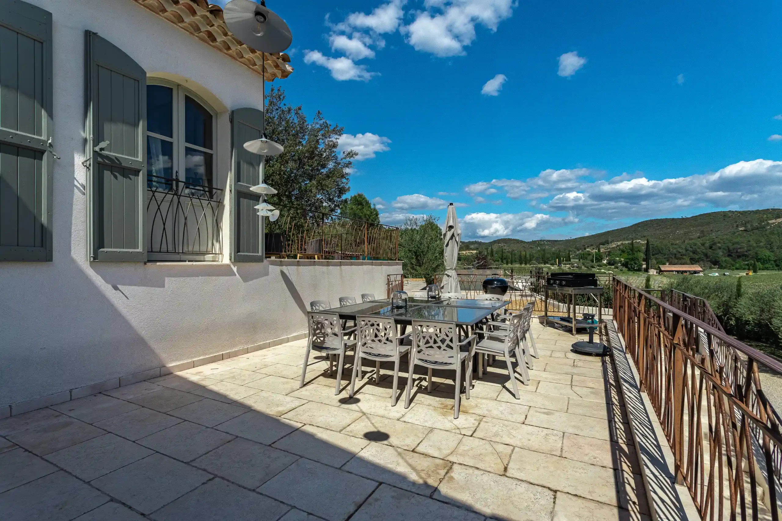 Terrasse avec grande table en verre, volets verts et vue sur les vignobles et collines