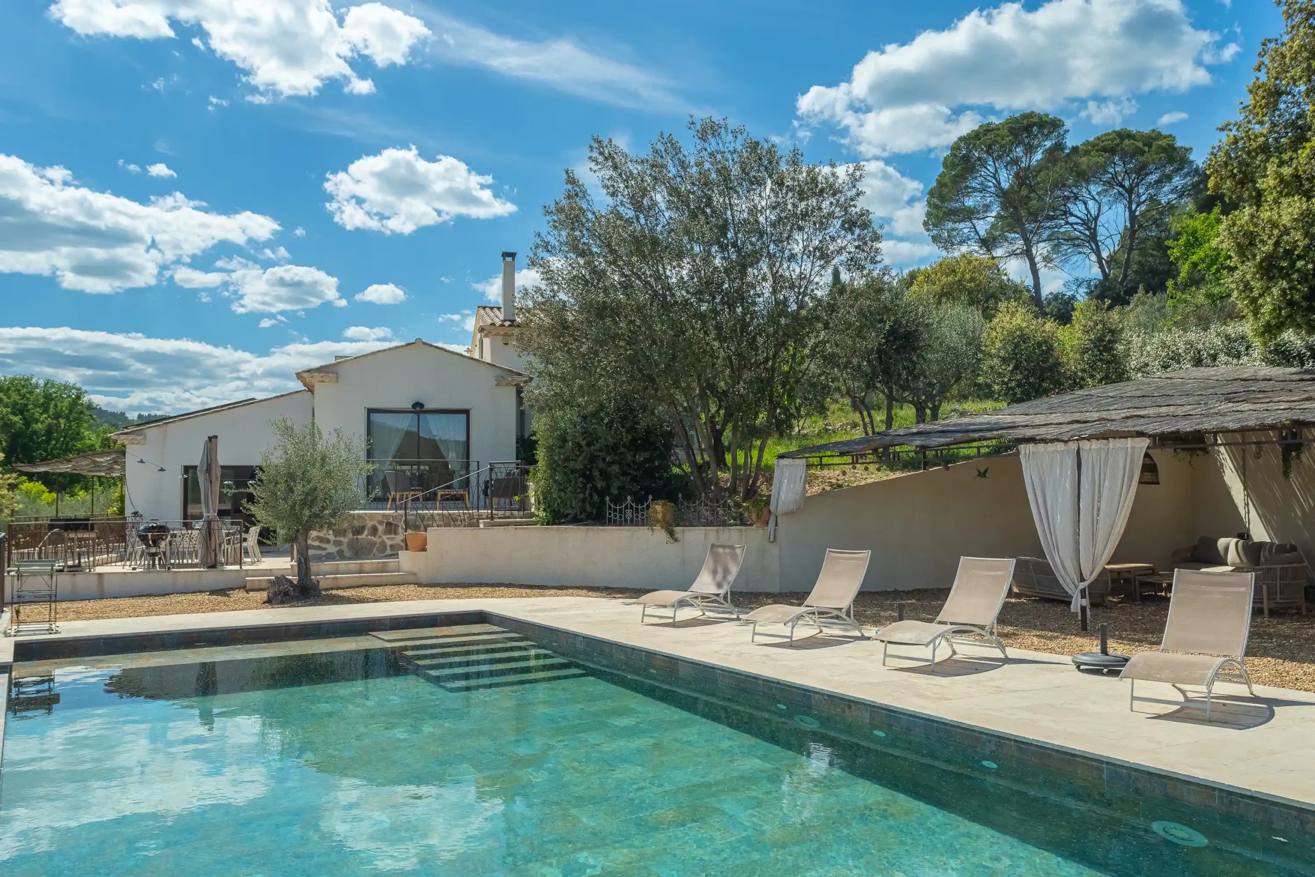 Piscine turquoise avec transats beiges, pergola aux rideaux blancs et grands oliviers