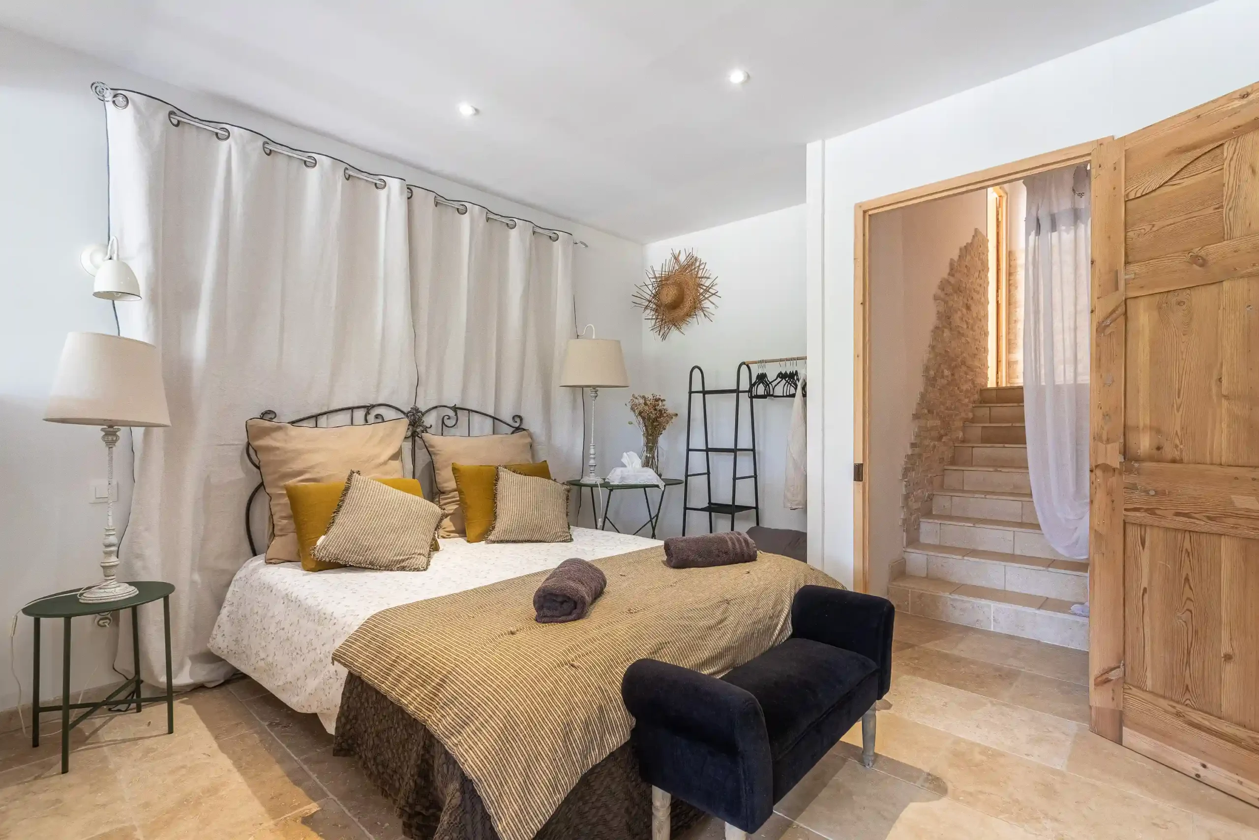 Chambre provençale avec grand lit en fer forgé, coussins ocres et porte en bois ouverte