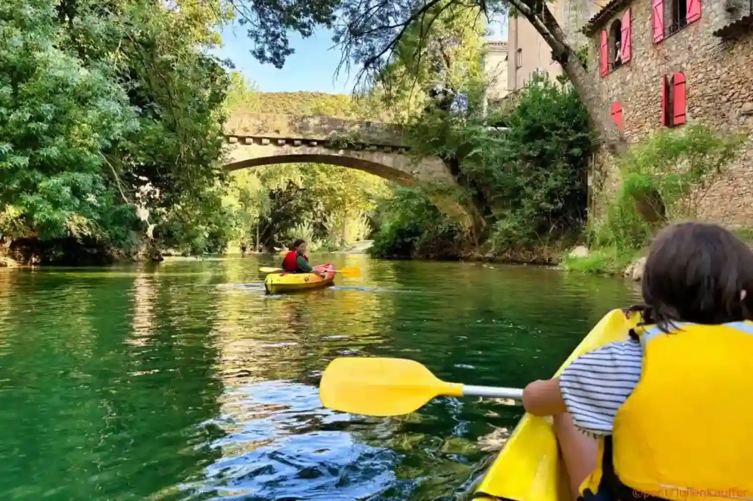 Enfant en gilet jaune pagayant en kayak sur une rivière verte près d'un pont en pierre